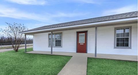 A small white house with a red door and a brown walkway leading to it.