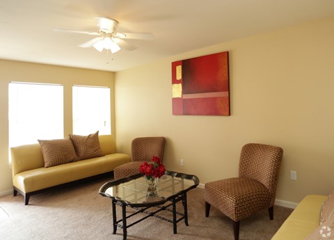 A living room with a yellow couch, a glass coffee table, and a red painting on the wall.