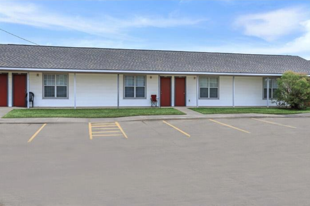 A white building with red doors and windows is surrounded by a parking lot.