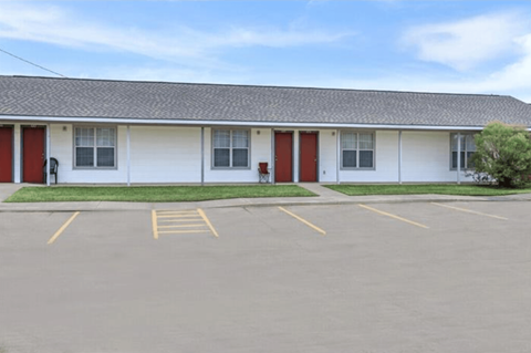 A white building with red doors and windows is surrounded by a parking lot.