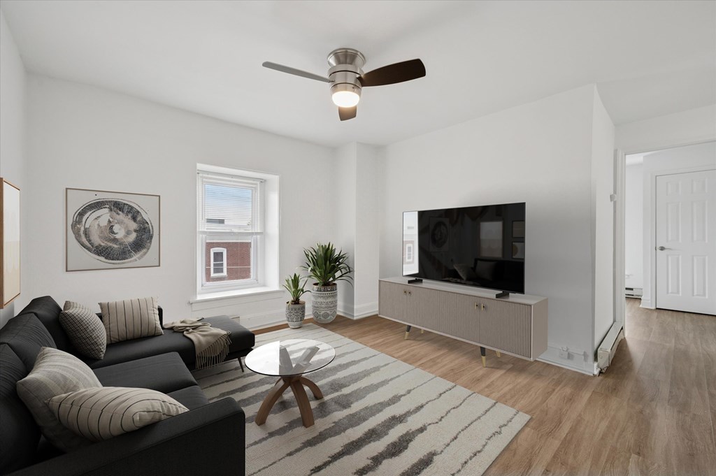 A living room with a black couch, a coffee table, a ceiling fan, and a television.
