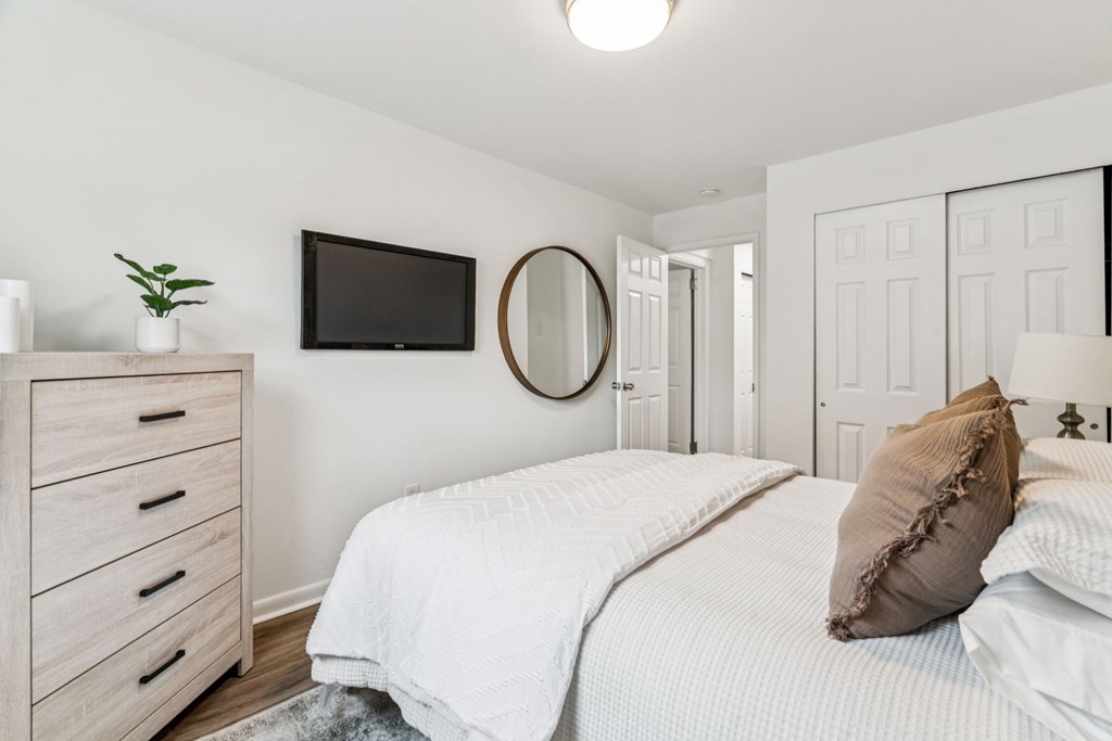 A white bedroom with a bed, dresser, mirror, and television.