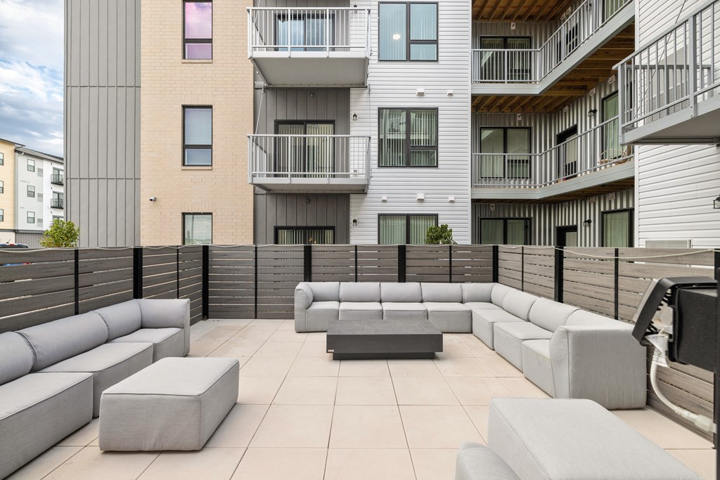 A modern outdoor seating area with grey couches and tables is surrounded by apartment buildings.