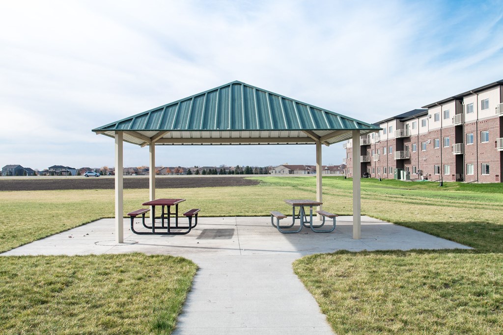 A green canopy pavilion with picnic tables in front of apartment buildings.