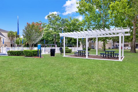 A white pergola with benches is situated in a grassy area.