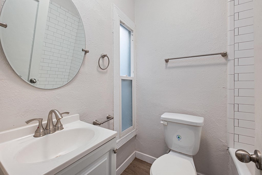 A white toilet with a blue sticker on the lid is in a bathroom with a white sink and mirror at Hillstone Flats Apartments, Los Angeles, CA