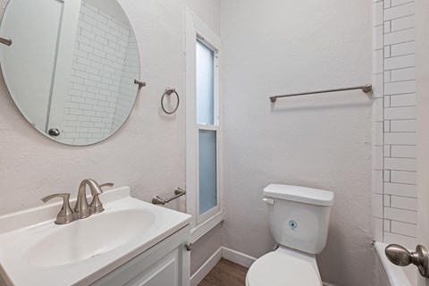 A white toilet with a blue sticker on the lid is in a bathroom with a white sink and mirror at Hillstone Flats Apartments, Los Angeles, CA