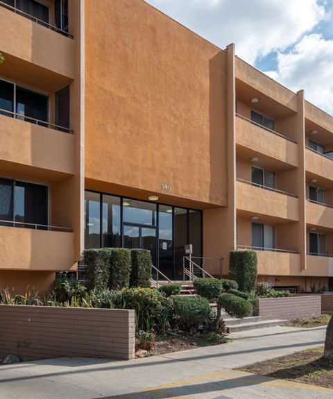 A modern apartment building with a brown facade and green bushes in front at Menlo Terrace Apartments, Los Angeles, CA