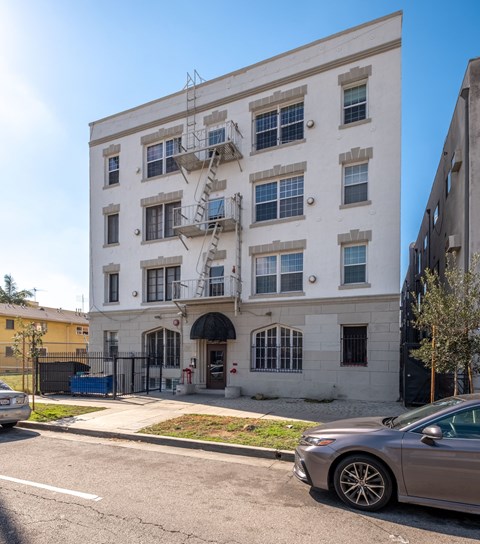 A silver car is parked on the side of a street in front of a white building with a fire escape at Normandie Summit Apartments, Los Angeles, CA