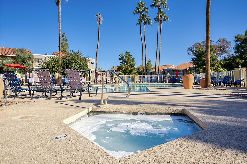 A hot tub sits in the middle of a patio with chairs and palm trees in the background.
