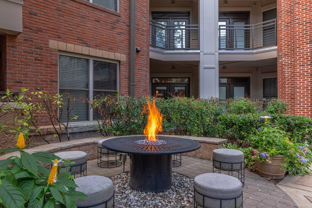 A fire pit sits in the middle of a courtyard surrounded by grey cushioned stools.