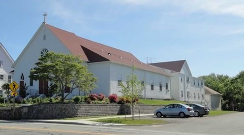 A church with a cross on top is surrounded by a stone wall and has a car parked in front.