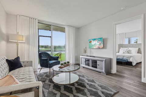 A living room with a grey sofa, a glass coffee table, a TV, and a window with curtains.
