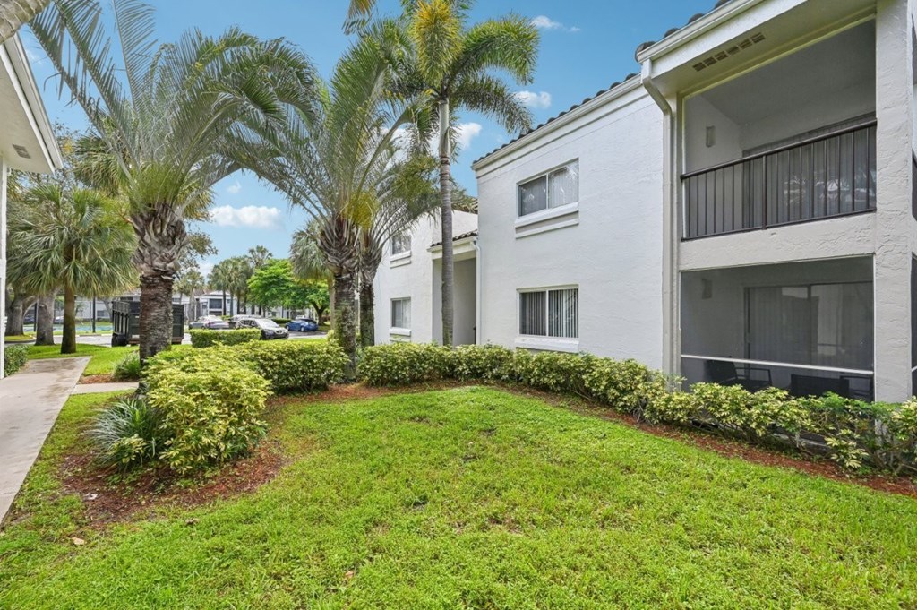 A white building with a balcony is surrounded by greenery and palm trees.