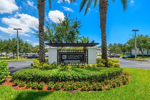 A sign for Legacy at Riverwalk Apartments is surrounded by greenery and palm trees.