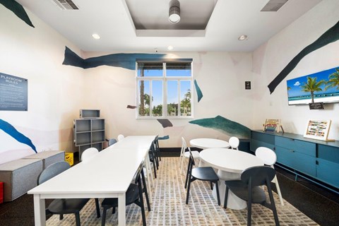 A room with a white table and chairs, a window with a view of trees, and a painting of a beach scene on the wall.