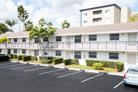 A parking lot in front of a white apartment building with a black car and a silver car parked.
