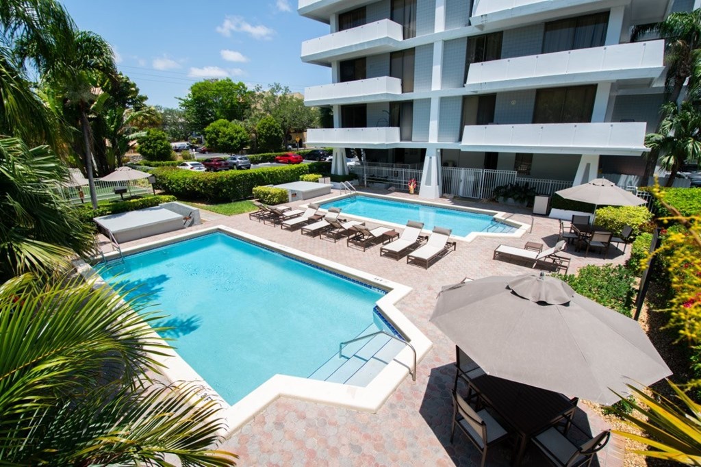 A large swimming pool surrounded by lounge chairs and umbrellas in front of a white apartment building.