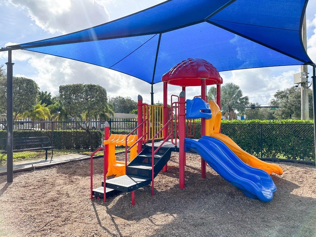 a playground with a slide and chairs under a blue umbrella