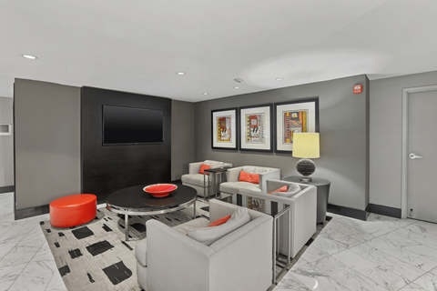 A modern living room with a grey couch, a black and white rug, a red ottoman, a black coffee table, a flat screen TV, and framed pictures on the wall.