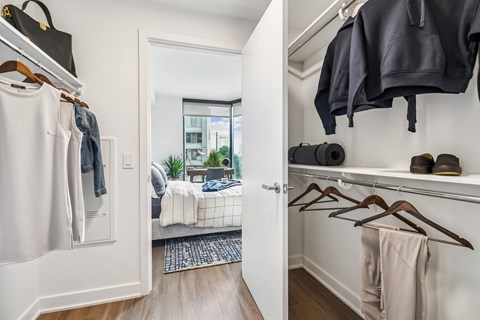 A white closet with clothes hanging and a bedroom in the background.