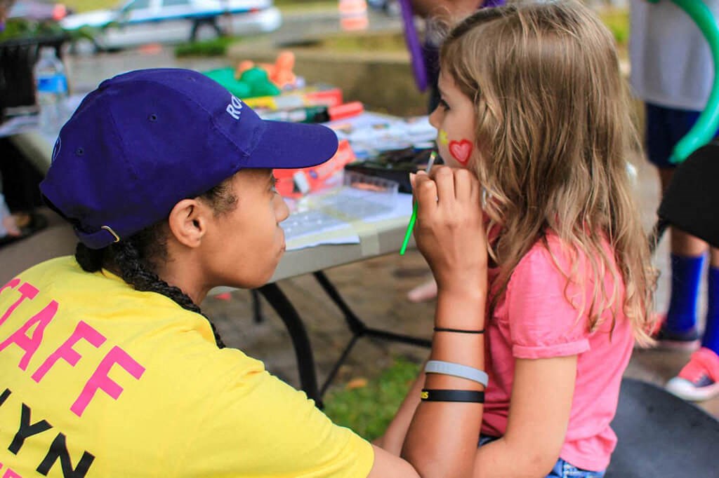 a little girl getting her face painted by a child at a picnic table