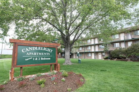 A sign for Candlelight Apartments stands in front of a tree and apartment building.