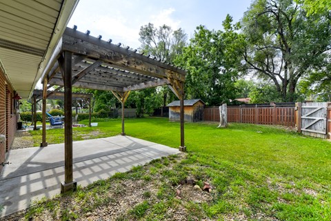A patio with a pergola and a carport is surrounded by a grassy area and trees.