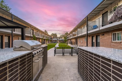 A sunset view of a courtyard with a BBQ grill and brick buildings.