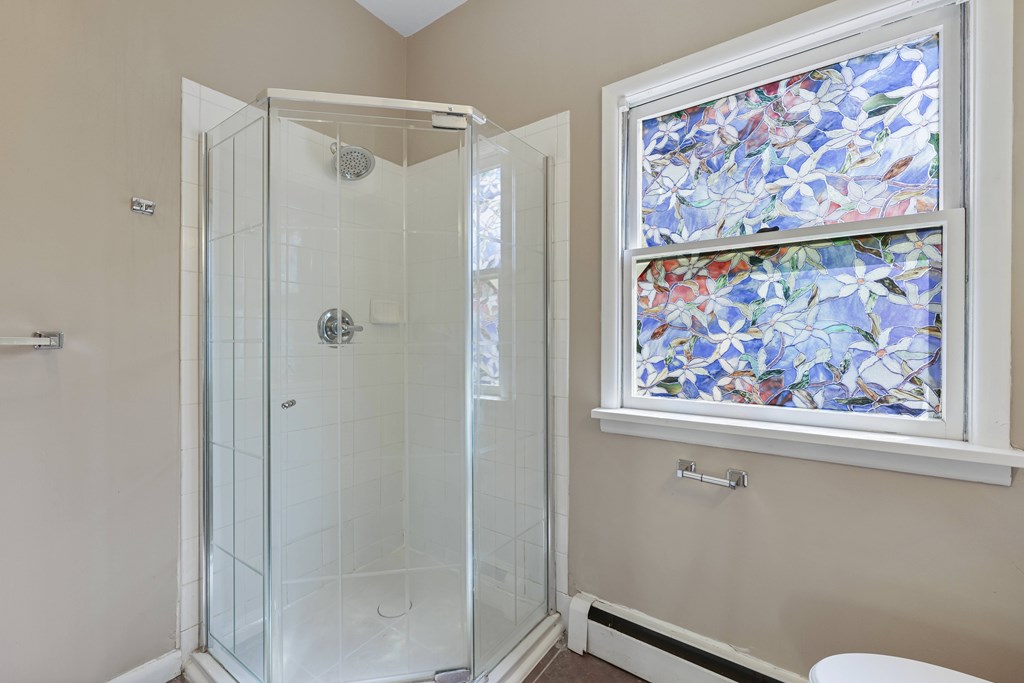 A shower with a glass door is in a bathroom with a window above it.