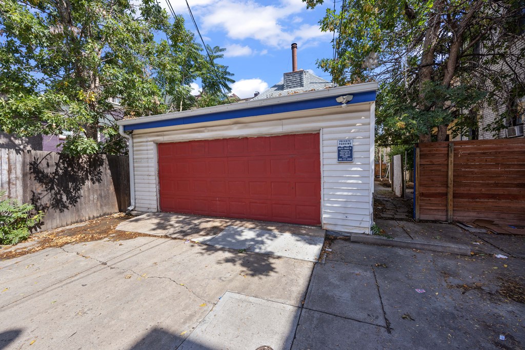 A small white building with a red garage door is surrounded by trees and a wooden fence.