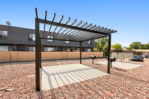 A pergola with a metal roof and wooden slats is situated in a gravel area with a building in the background.