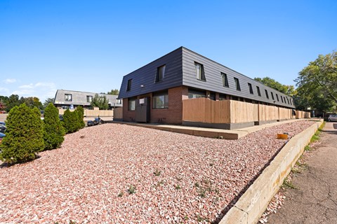 A modern building with a grey roof and wooden exterior is surrounded by a gravel area.