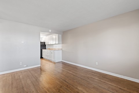 A kitchen with white cabinets and a black fridge is visible through the open door of a room with wooden floors.