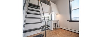 A staircase with a metal railing is in the foreground of a room with a kitchen in the background.