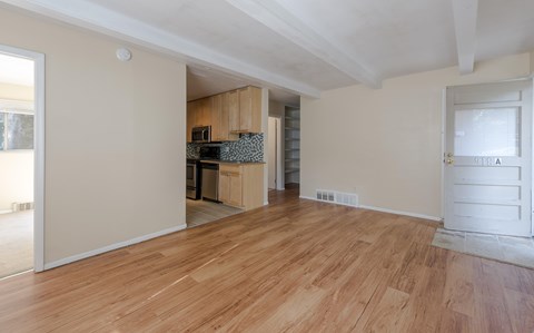 A room with wooden flooring and a kitchen area in the background.