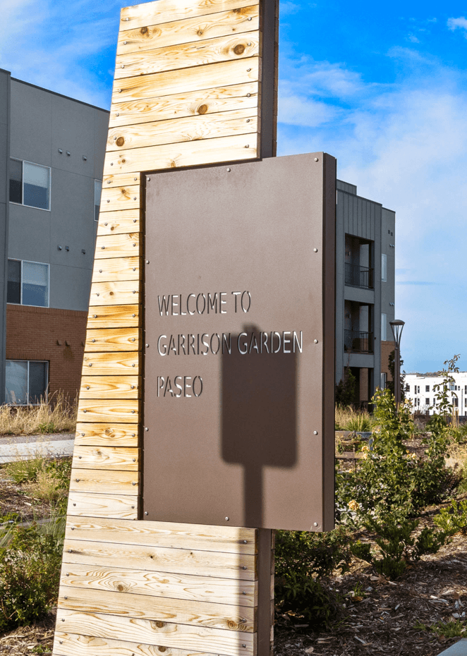 A brown sign reads "Welcome to Garrison Garden Paseo" in front of a building.