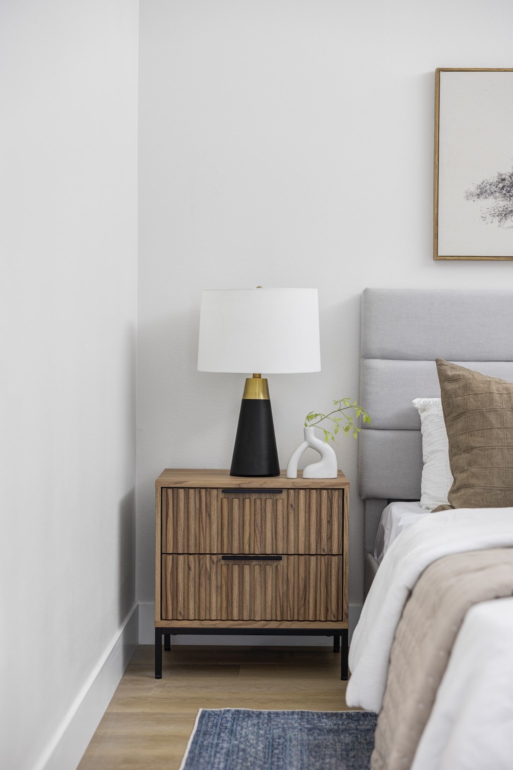 A lamp with a black base and a white lampshade is on a wooden cabinet next to a bed with a grey headboard.