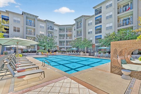 A large swimming pool surrounded by lounge chairs and umbrellas in front of apartment buildings.