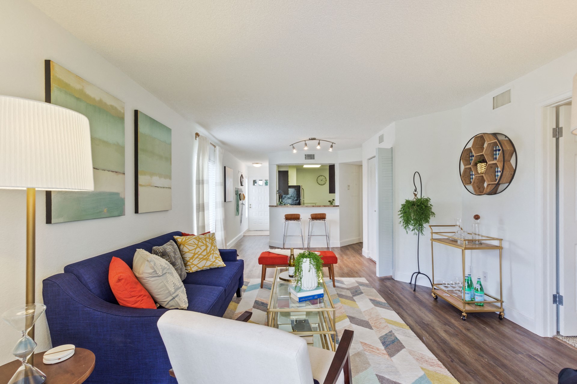 Living room with patterned area rug and colorful couch, chair, and ottoman with coffee table looking into kitchen with bar and two stools