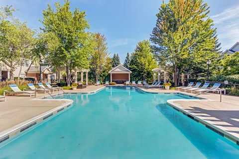 Apartments in Aurora, IL, for Rent - The Apartments at Kirkland Crossing - A Large Sparkling Swimming Pool Surrounded by Tanning Chairs, Tall Trees, and Lush Greenery.