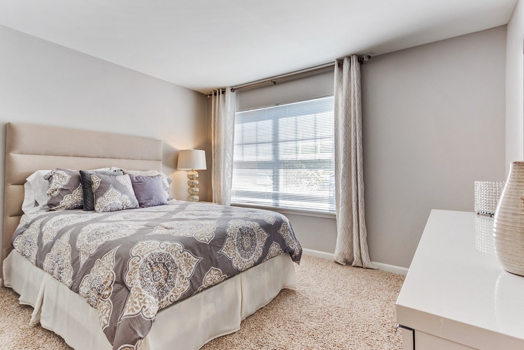 Apartments In Aurora, IL - The Apartments At Kirkland Crossing - Bedroom With Bed, Dresser, Carpet Flooring, Nightstand, Lamp, And Window.