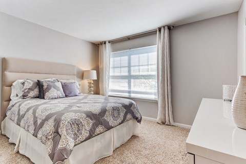 One-Bedroom Apartments in Aurora, IL - The Apartments at Kirkland Crossing - Spacious Bedroom with Plush Carpeting, Lamp, and a Large Window with Curtains and Blinds.