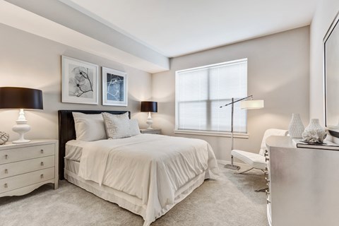 Apartments for Rent in Vernon Hills, IL - The Oaks of Vernon Hills - Cozy Bedroom With Plush White Bedding, Framed Wall Art, Soft Carpet, Dual Nightstands, and Wide Window With Blinds.