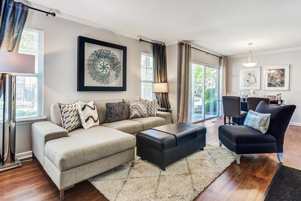 Aurora, IL Apartments For Rent - The Apartments At Kirkland Crossing - Living Room With Sectional, Chair, Ottoman, Wood-Style Flooring, Modern Decor, Area Rug, Sliding Glass Door To Back, And Floor Lamps.