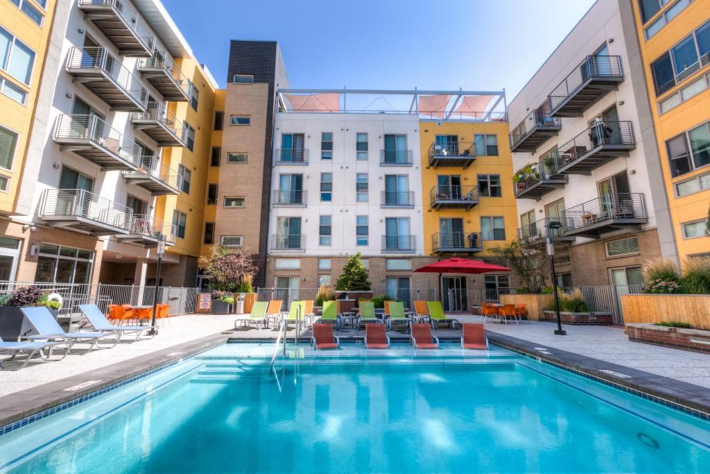 1 BR Apartments in Denver CO - Line 28 LoHi - Spacious Bedroom with Carpeted Floor, Bed, Wooden Nightstands with Lamps and Dresser, Standing Lamp, Ceiling Fan, and Window with Curtains