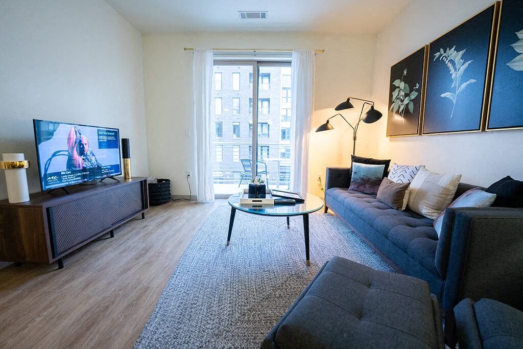 Denver CO Apartments - Line 28 LoHi - Furnished Living Room with Sofa, Chair and Ottoman, Glass Coffee Table, TV Stand with TV, Balcony Access, Large Windows with Curtain, and Plank Wood Floor