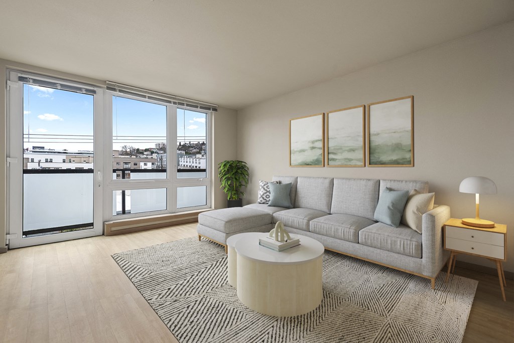 A living room with a grey couch, a coffee table, and a large window overlooking a cityscape.