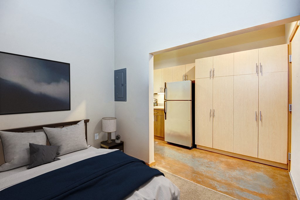 A bedroom with a bed, a painting, a lamp, a refrigerator, and a cabinet.