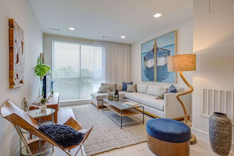 A living room with a white couch, a blue ottoman, and a large painting on the wall.